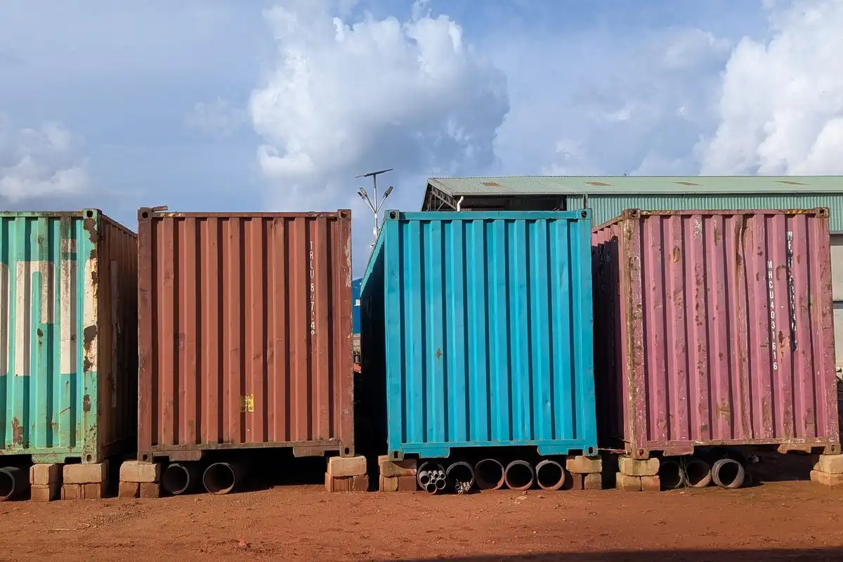 Four shipping containers in green, rust, blue and red standing on red earth under a cloudy sky.