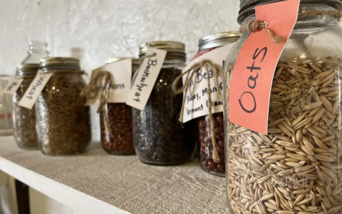 Glass jars of oats, lentils and other grains on a shelf, each with a handwritten paper tag.
