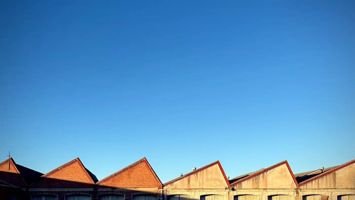 A row of brick warehouse roofs with sawtooth gables under a clear blue sky.