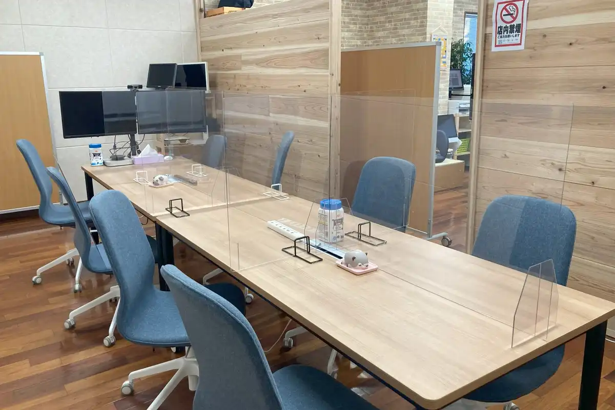 A shared work table with blue chairs, monitors and clear desk screens in a bright office.