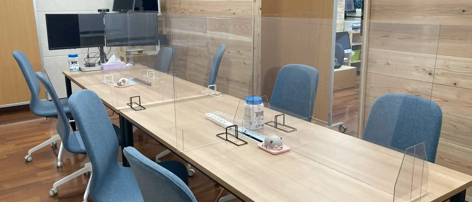 A shared work table with blue chairs, monitors and clear desk screens in a bright office.