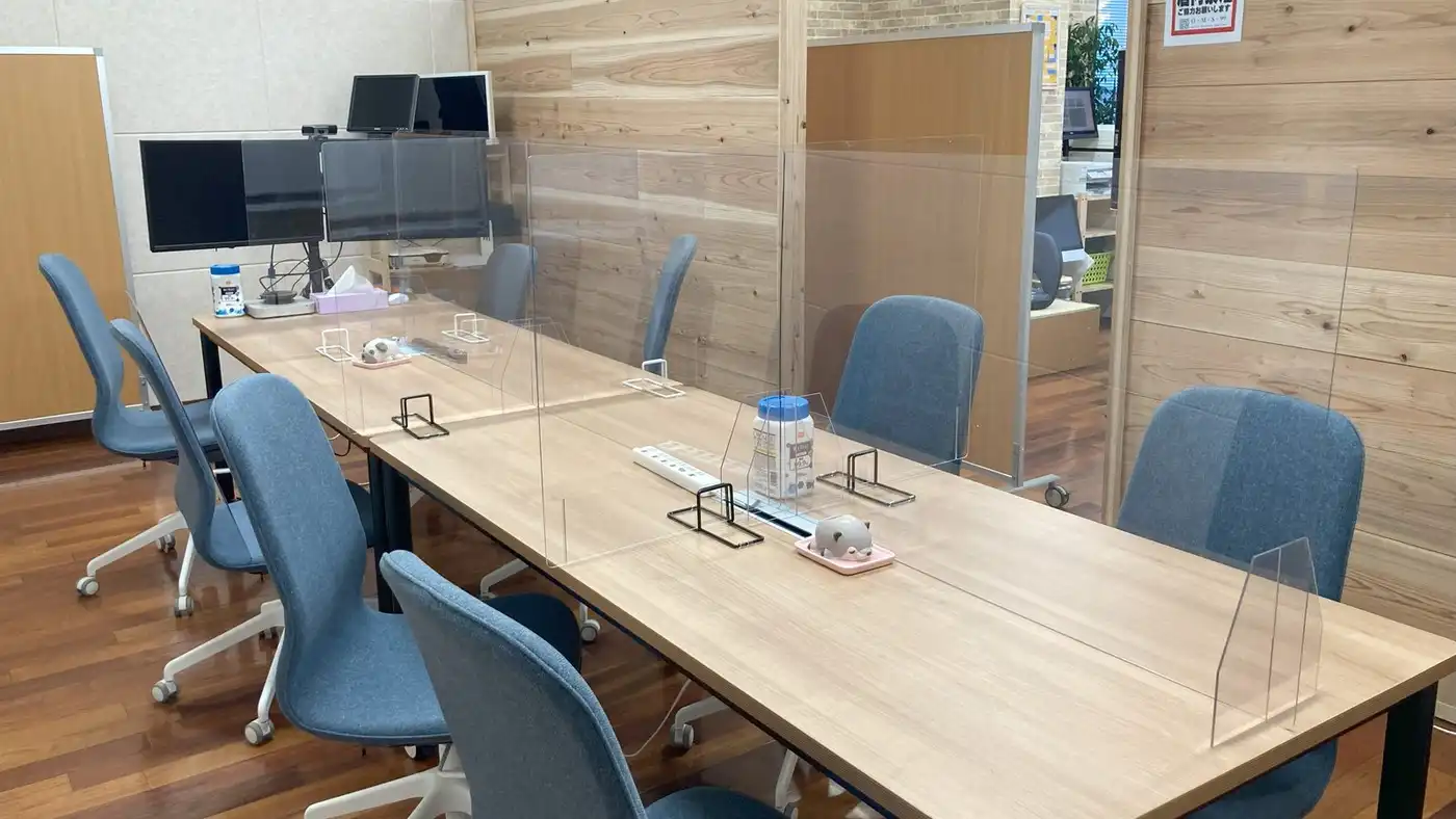 A shared work table with blue chairs, monitors and clear desk screens in a bright office.